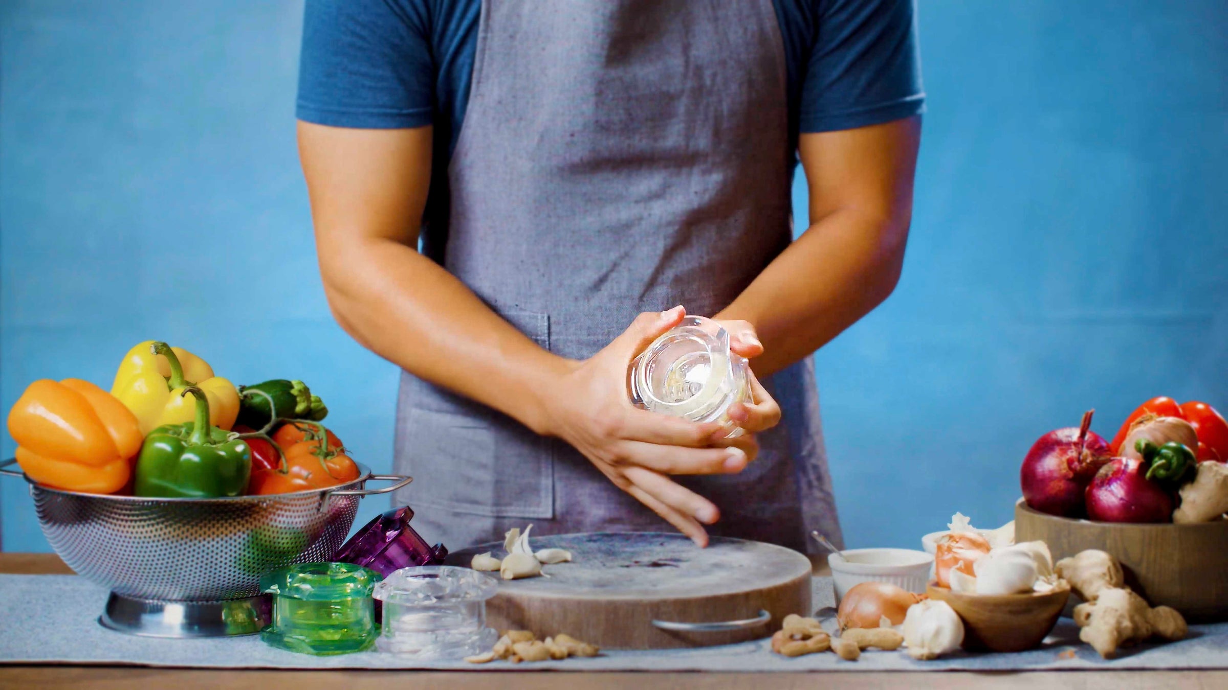 Man standing with a garlic twister in his hands, demonstrating the twisting motion. Other ingredients including onion, peppers and shallots are shown.